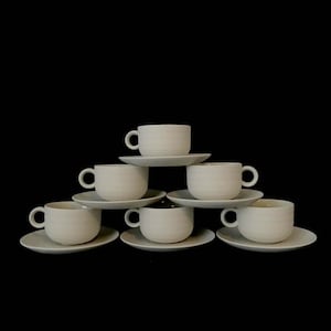 May include: A set of six white ceramic teacups and saucers stacked in a pyramid shape. The cups and saucers are plain white with no decorations.