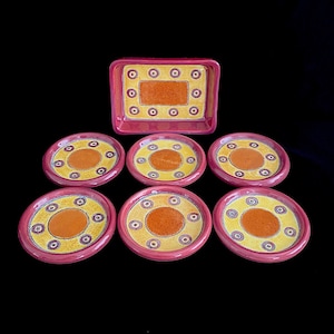 May include: Set of seven ceramic coasters with a yellow and orange geometric design. The coasters have a red rim and are arranged in a pyramid shape.