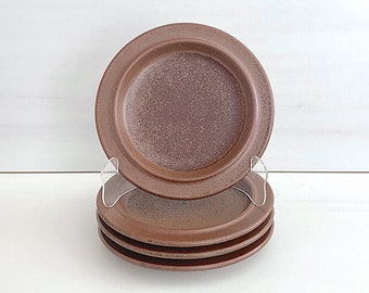May include: Set of four brown ceramic saucer plates. The plates have a speckled glaze and a slightly raised rim.