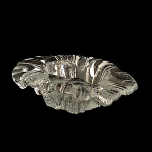 May include: A clear glass ashtray with a wavy, organic design. The ashtray is made of thick glass and has a slightly rippled surface.