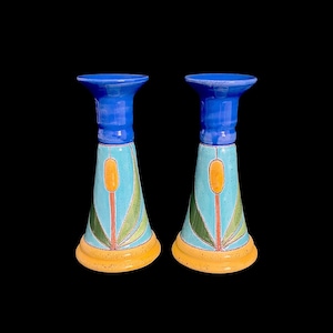 May include: Two ceramic candle holders with a blue top and base, a turquoise middle section, and an orange and green floral design. The base is a speckled yellow. The candle holders are on a black background.