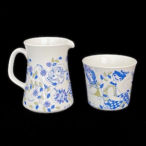 May include: A white ceramic pitcher and a white ceramic cup with a blue and green floral pattern featuring stylized figures.