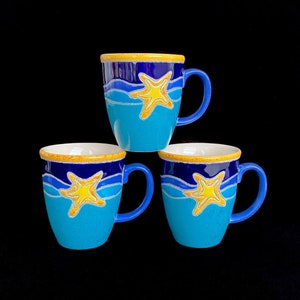 May include: Three blue ceramic mugs with yellow and blue starfish designs. The mugs have white rims and handles.