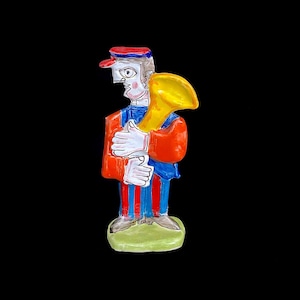 May include: A ceramic figurine of a person wearing a red and blue striped outfit and a red hat, holding a yellow trumpet.