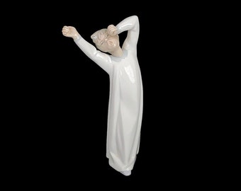 May include: A white porcelain figurine of a person in a white nightgown, stretching with one arm raised and the other hand covering their face.