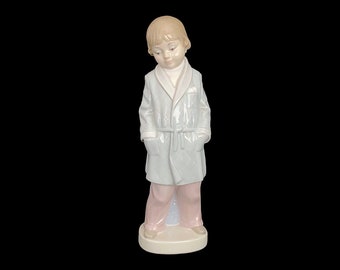 May include: A porcelain figurine of a young boy wearing a light blue robe and pink pants. The figurine is standing on a cream-colored base.