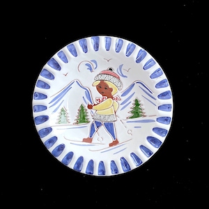 Puede incluir: Un plato de cerámica blanco con un borde azul y blanco. El plato presenta una ilustración pintada a mano de una persona esquiando en un paisaje invernal con árboles y montañas.