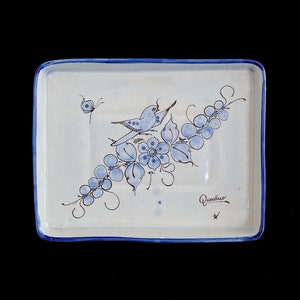 May include: A white ceramic square dish with a blue rim and a blue and white floral design featuring a bird, grapes, and leaves. The dish is signed "Quenelano" in the bottom right corner.