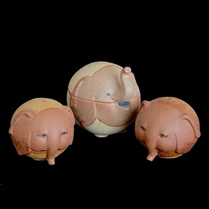 May include: Three ceramic elephant salt and pepper shakers. The shakers are brown and beige with painted details. The middle shaker is larger and has a lid.