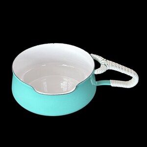 May include: A round, turquoise enameled metal bowl with a smooth white interior and a single side handle wrapped in white woven material. The bowl features a small pouring spout on the rim, providing a functional design for kitchen or serving use.