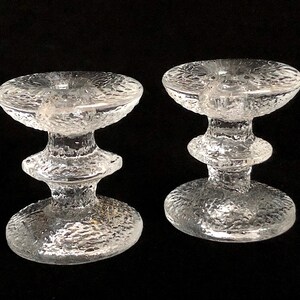 May include: Two clear glass candle holders with a textured surface. The holders have a wide base and a narrow middle section.