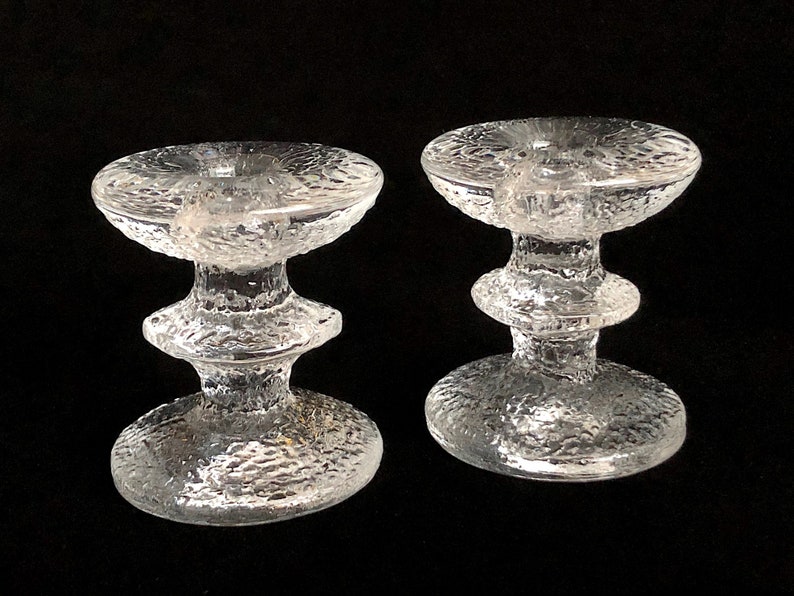 May include: Two clear glass candle holders with a textured surface. The holders have a wide base and a narrow middle section.