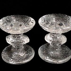 May include: A pair of clear glass candle holders with a textured surface. Each holder has a wide, round base and a three-tiered stem.