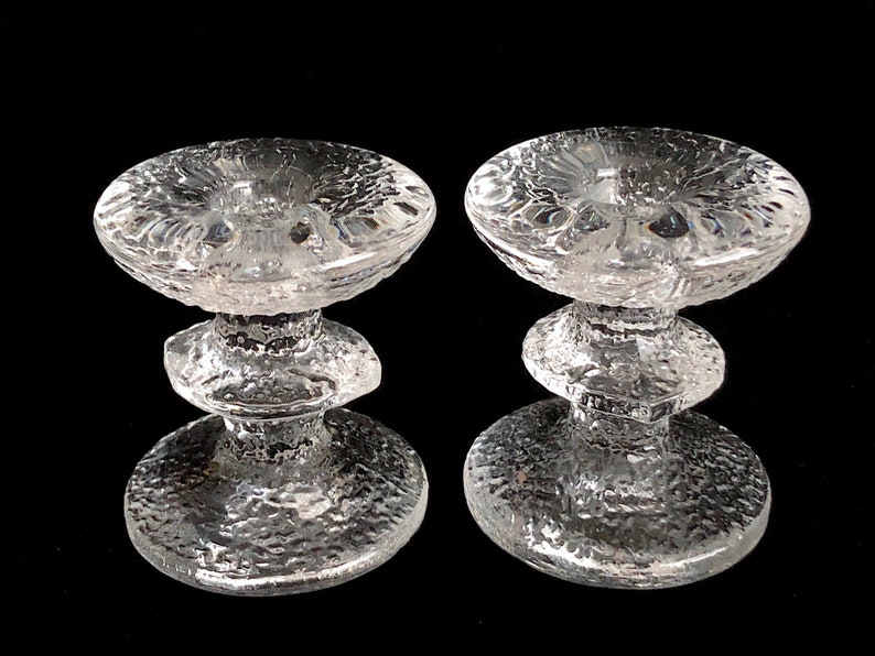May include: A pair of clear glass candle holders with a textured surface. Each holder has a wide, round base and a three-tiered stem.