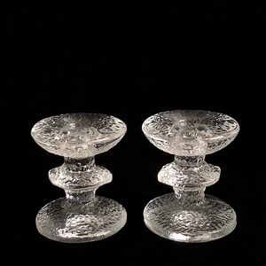 May include: Two clear glass candle holders with a textured surface. Each holder has a wide base and a narrow middle section.