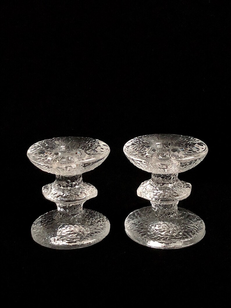 May include: Two clear glass candle holders with a textured surface. Each holder has a wide base and a narrow middle section.