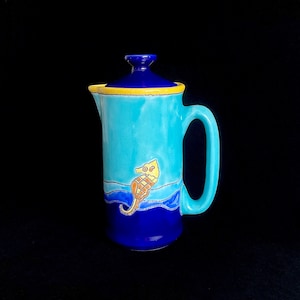 May include: A blue and yellow ceramic pitcher with a seahorse design. The pitcher has a blue lid and a handle.