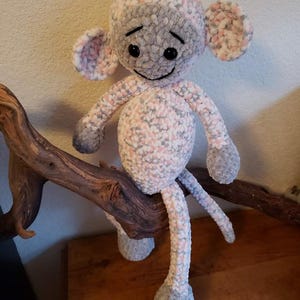 Puede incluir: Un muñeco de mono de ganchillo hecho a mano en tonos grises, rosas y blancos. El peluche tiene orejas grandes, una cara sonriente y brazos y piernas largas. El mono está sentado en una rama marrón, con una superficie de madera en el fondo.