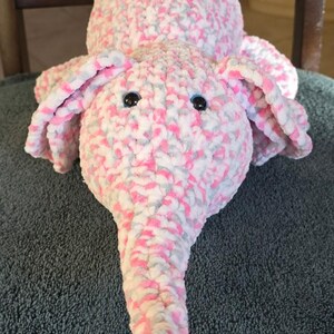 Puede incluir: Un juguete de peluche de elefante hecho a mano en tonos de rosa, blanco y gris. El elefante tiene orejas grandes y caídas, una larga trompa y pequeños ojos negros. La tela suave y texturizada le da al juguete una apariencia tierna.
