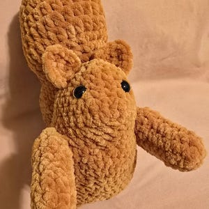 May include: A plush, handmade squirrel toy in shades of brown. The stuffed animal has a fluffy, textured body, small rounded ears, and black button eyes. The squirrel's tail is curled over its back, and its arms are outstretched.
