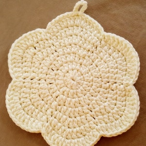 Puede incluir: Salvamanteles de ganchillo color crema con forma de flor. El salvamanteles está hecho de hilo grueso y tiene un lazo para colgar. El diseño presenta una serie de círculos concéntricos, creando una superficie texturizada. La estética general es artesanal y rústica.