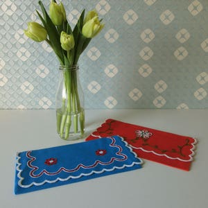 May include: Two embroidered fabric napkins, one blue and one red, with white scalloped edges and floral designs. The napkins are on a white surface.