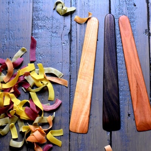 Könnte beinhalten: Drei Holzschuhspanner in verschiedenen Farben: Naturholz, dunkelbraunes Holz und rötlich braunes Holz. Die Schuhspanner sind vertikal auf einer Holzoberfläche angeordnet.