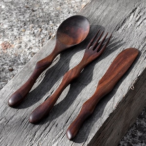 Puede incluir: Un juego de tres utensilios de madera, una cuchara, un tenedor y un cuchillo, hechos de madera marrón oscura. Los utensilios tienen mangos únicos y curvos.