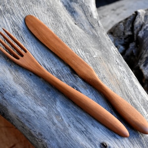 Può includere: Un set di posate in legno, composto da una forchetta e un coltello, entrambi con una finitura in legno naturale. Le posate sono disposte su un pezzo di legno invecchiato.