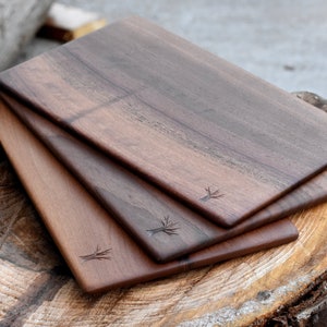 May include: Three wooden cutting boards made of walnut wood. The boards are stacked on top of each other and have a natural wood finish. Each board has a small tree design carved into the surface.
