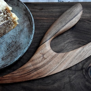 Puede incluir: Un cuchillo para pastel de madera con un diseño curvo único. El cuchillo está hecho de madera oscura con un patrón de veta de madera más claro. El cuchillo tiene un pequeño símbolo grabado en el mango.