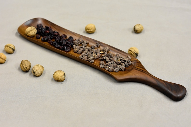 May include: A long, wooden serving tray with a handle. The tray is filled with dried cranberries and candied nuts.