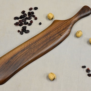 May include: A long, dark brown wooden serving paddle with a curved handle. The paddle is made of walnut wood and has a natural finish.