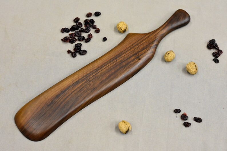 May include: A long, dark brown wooden serving paddle with a curved handle. The paddle is made of walnut wood and has a natural finish.