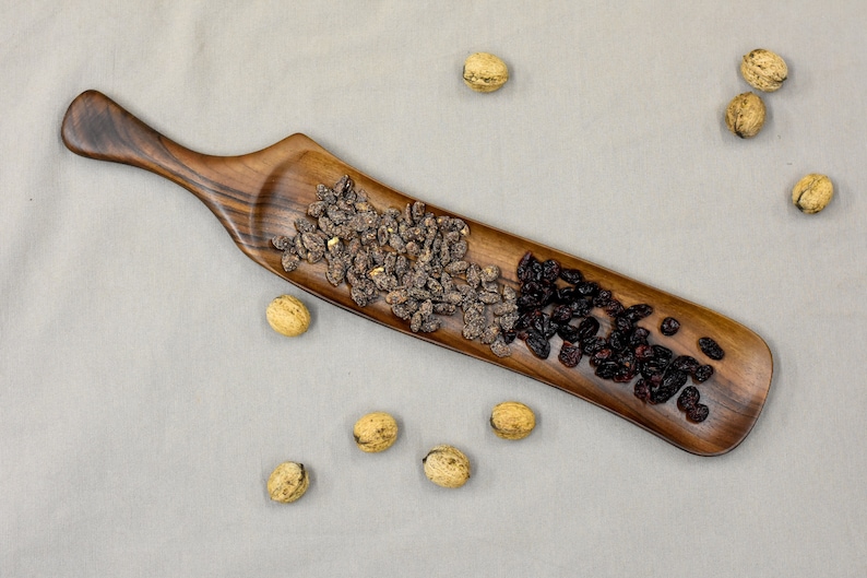 May include: A long, wooden serving tray with a curved handle. The tray is filled with two types of dried fruit: dark brown and black. The tray is on a light gray surface with several walnuts scattered around it.
