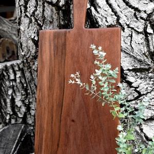 May include: A large, rectangular wooden cutting board with a handle. The board is made of dark wood with a natural grain pattern. The board is sitting on a piece of tree bark.