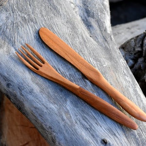 May include: A wooden fork and knife set on a weathered wooden surface. The utensils are made of natural wood and have a simple, rustic design.