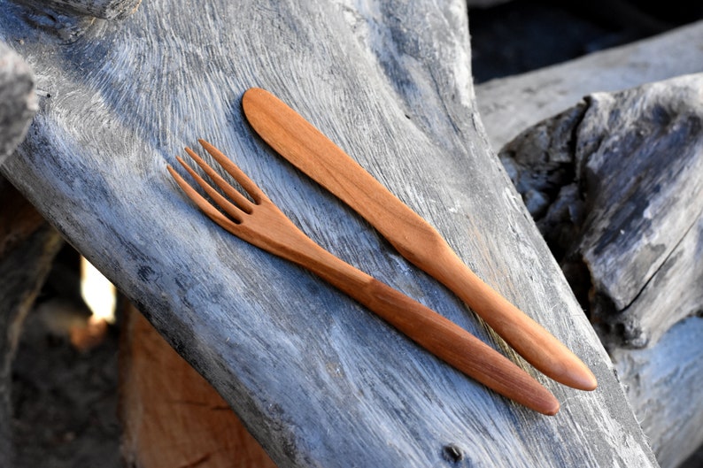 May include: A wooden fork and knife set on a weathered wooden surface. The utensils are made of natural wood and have a simple, rustic design.