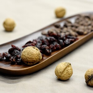 May include: A wooden tray filled with dried cranberries, chocolate chips, and walnuts. The tray is on a light brown fabric surface.