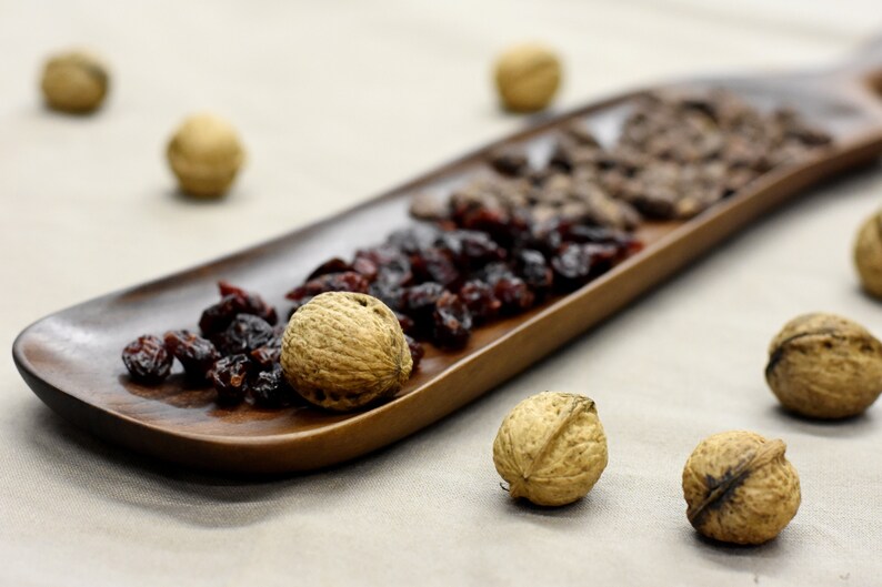 May include: A wooden tray filled with dried cranberries, chocolate chips, and walnuts. The tray is on a light brown fabric surface.