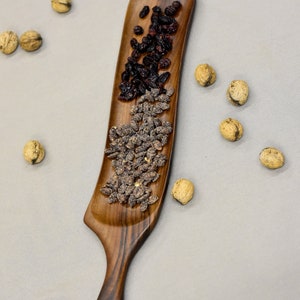 May include: A wooden serving tray with a long, narrow handle. The tray is filled with dried cranberries and sunflower seeds.
