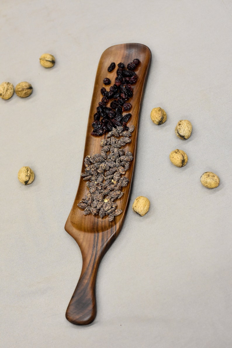 May include: A wooden serving tray with a long, narrow handle. The tray is filled with dried cranberries and sunflower seeds.