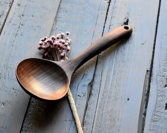 May include: A dark brown wooden serving spoon with a long handle. The spoon is lying on a light gray wooden surface with a small bouquet of dried flowers.