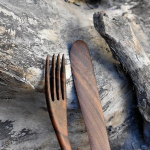 May include: A wooden fork and knife set, made from dark brown wood with a natural grain pattern. The utensils are resting on a piece of weathered driftwood.