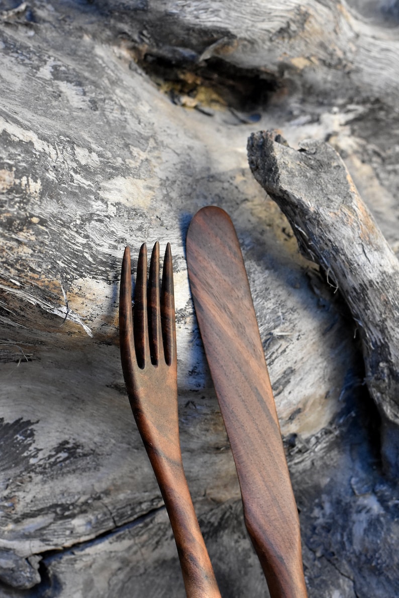 May include: A wooden fork and knife set, made from dark brown wood with a natural grain pattern. The utensils are resting on a piece of weathered driftwood.