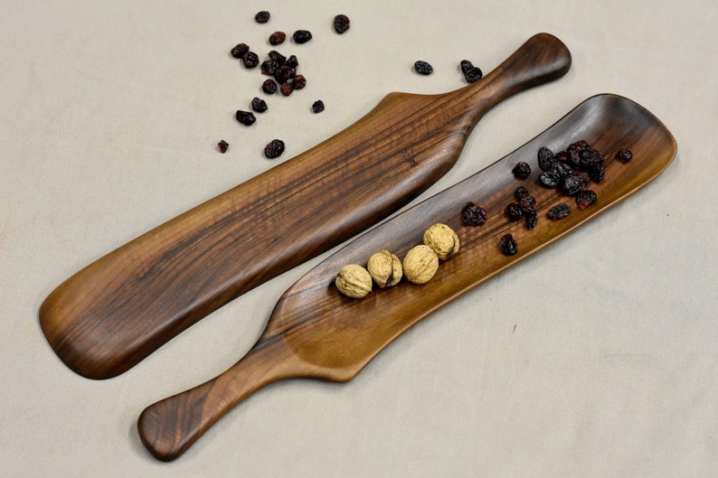 May include: Two wooden serving trays with handles. The larger tray is longer and narrower than the smaller tray. The smaller tray has a few walnuts and dried cranberries on it.