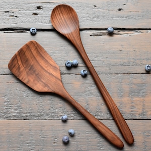 Może przedstawiać: Dwa drewniane narzędzia kuchenne, łyżka i szpatułka, leżą na rustykalnej drewnianej powierzchni z rozrzuconymi jagodami.
