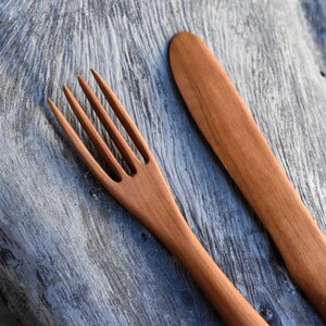 May include: A wooden fork and knife set, both with a natural brown finish. The utensils are lying on a gray wooden surface.