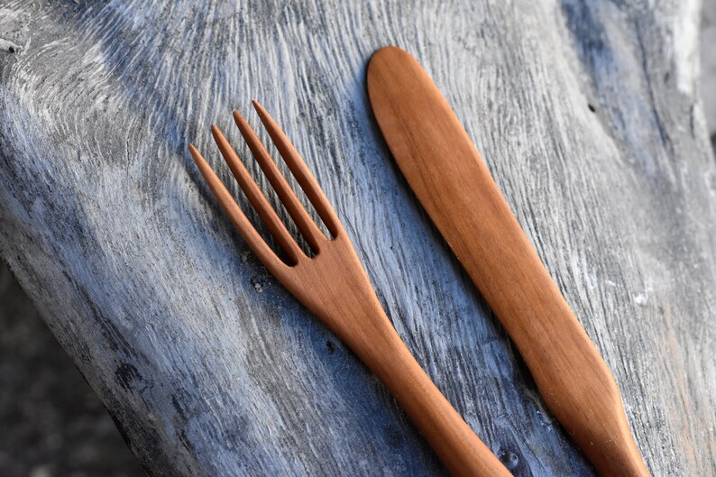 May include: A wooden fork and knife set, both with a natural brown finish. The utensils are lying on a gray wooden surface.