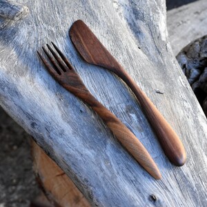 May include: A wooden fork and knife set made from dark brown walnut wood. The utensils are lying on a weathered gray wooden surface.
